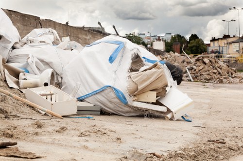 A large pile of mixed waste materials lies on a dirt surface in an open area, including white plastic pipes, a torn white and blue tarpaulin, and various other debris. The waste appears to be construction or renovation refuse, with some items partially folded or crumpled, and scattered across the ground. In the background, there is a partly demolished brick wall, along with distant trees, buildings, and utility poles, under a cloudy sky. The scene suggests a site undergoing construction or clearance, where rubbish removal services such as those offered by Commercial Waste Reigate are often required to manage and clear waste efficiently from commercial or construction environments in the Reigate area.