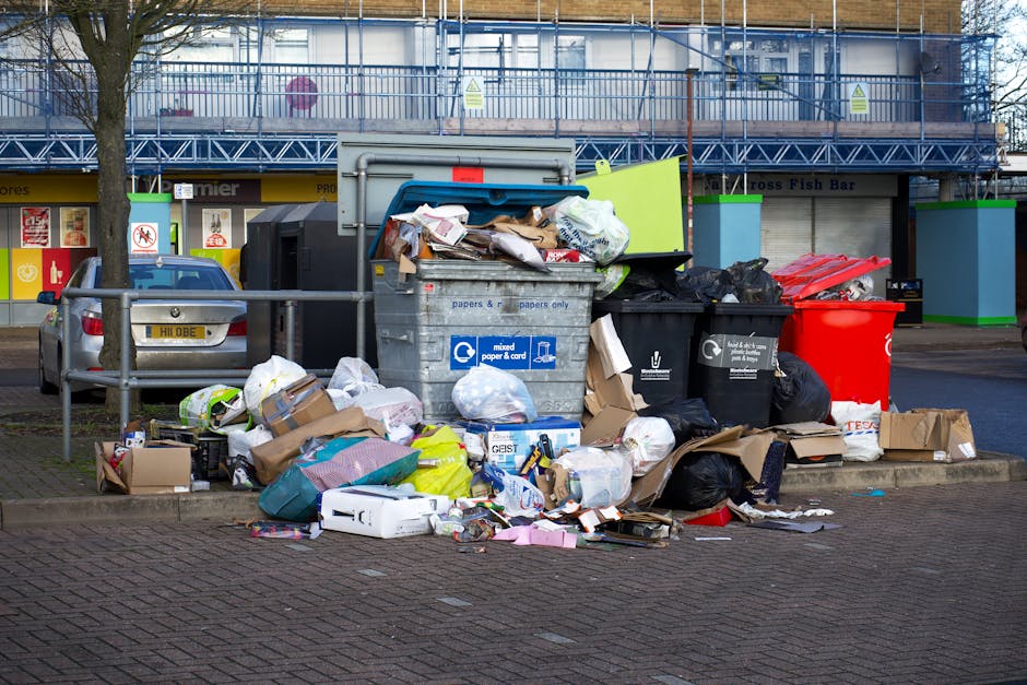 A middle-aged man with dark hair, wearing a black T-shirt with white graphic text on the front, is bent over, disposing of waste into a modern, stainless steel outdoor litter bin with a cylindrical sh