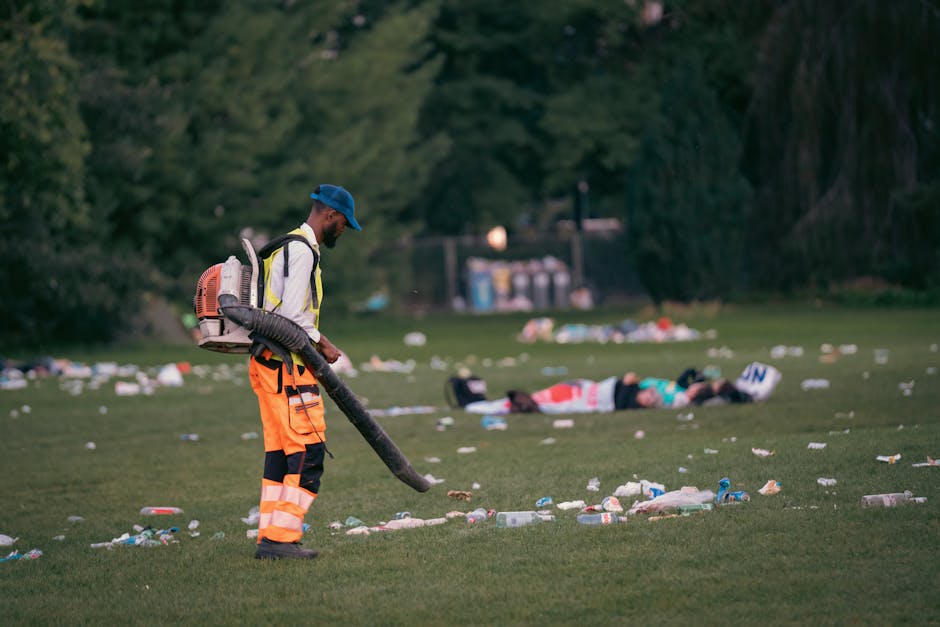 A tall, blue plastic rubbish bin with a closed lid, positioned outdoors on a grassy area near a paved pathway, is the primary object in the image. The bin features a rectangular shape with a slightly 
